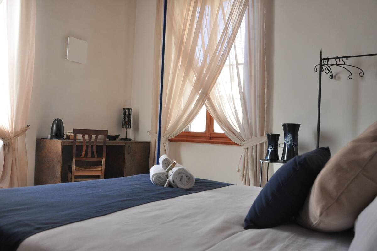 Un dormitorio con cama, escritorio, silla y ventana con cortinas traslúcidas.