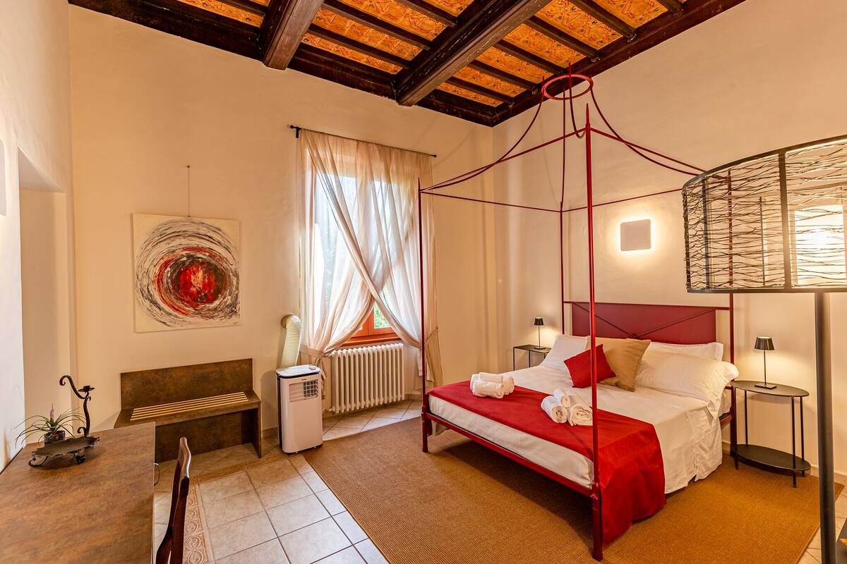 Un dormitorio con cama con dosel, un banco, un radiador, una mesita de noche y una obra de arte colgada en la pared.