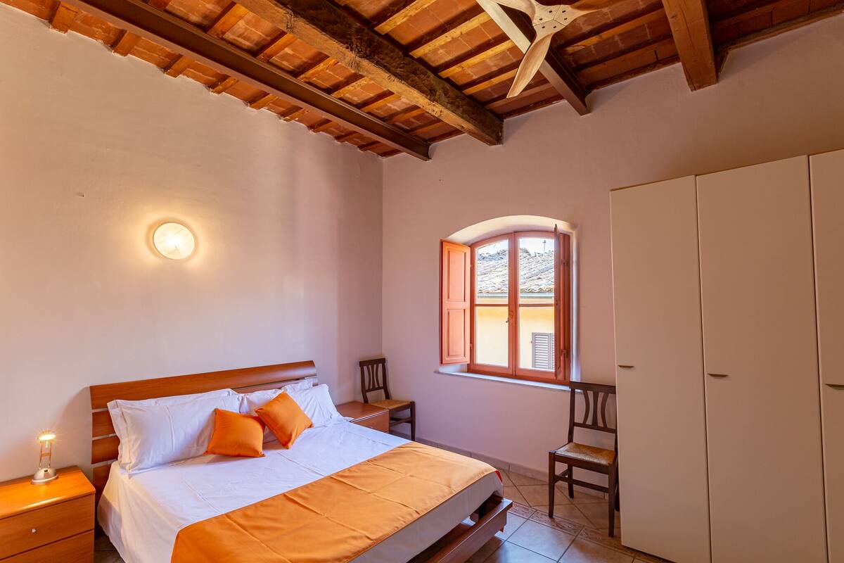 Un dormitorio con techo de madera, una cama con ropa de cama blanca y naranja, una mesita de noche con una lámpara, una silla, un armario y una ventana con vistas al exterior.