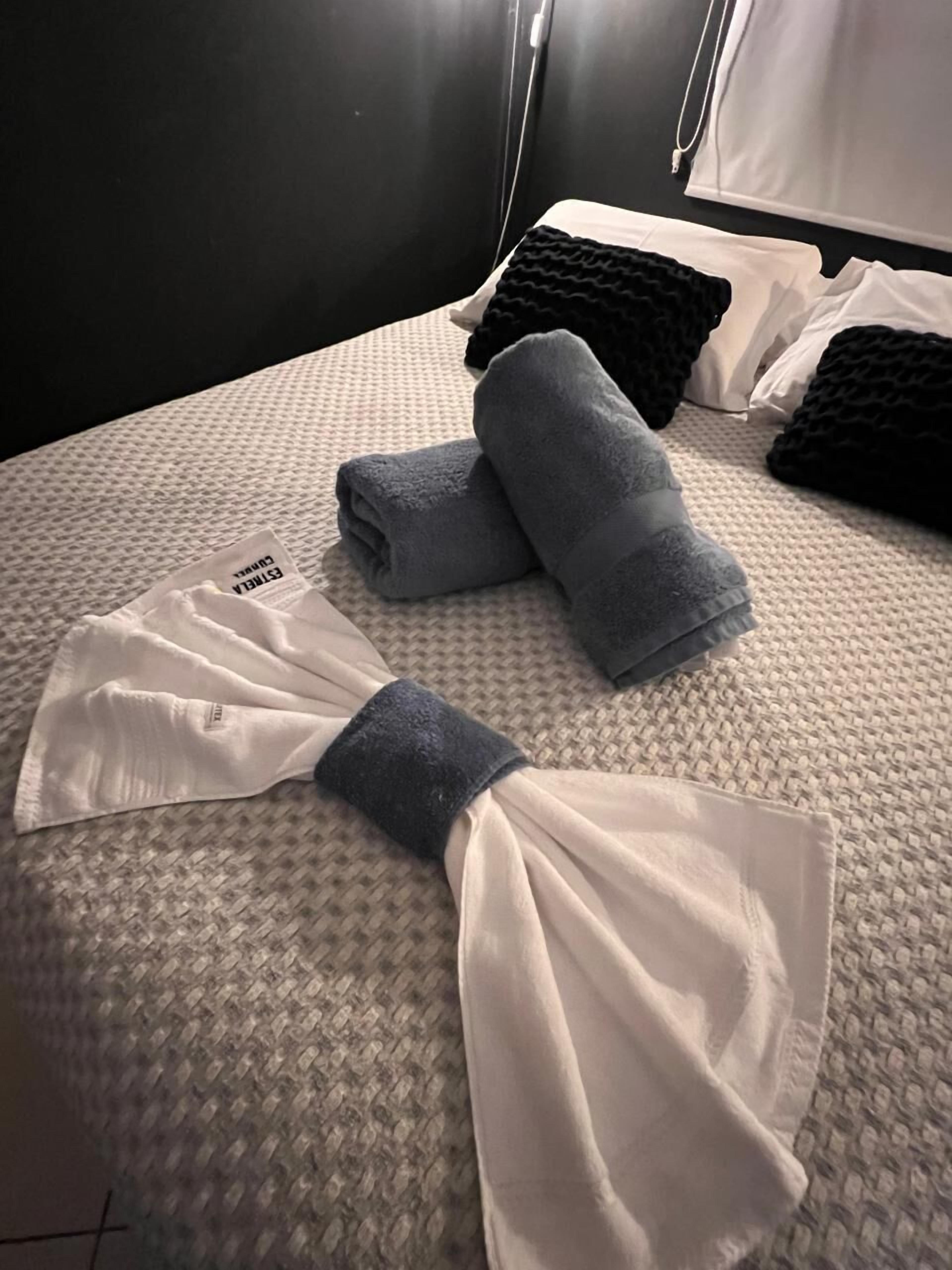 A bed with a textured grey bedspread, two rolled-up towels, and a white pillow.
