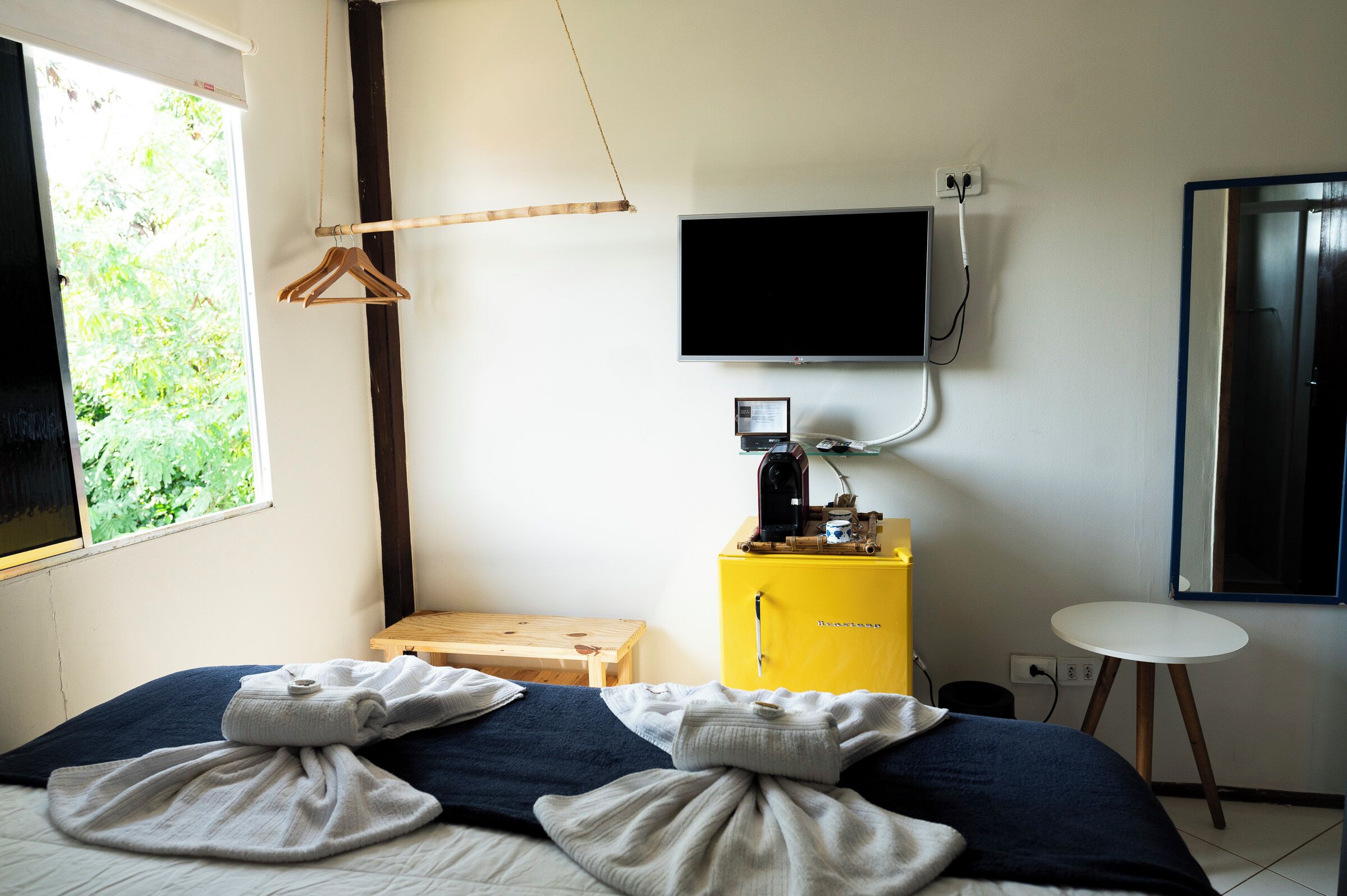 Ein Schlafzimmer mit einem Bett, einem kleinen Holztisch, einer gelben Minibar, einem Fernseher und einem Fenster mit Blick auf Grünflächen.