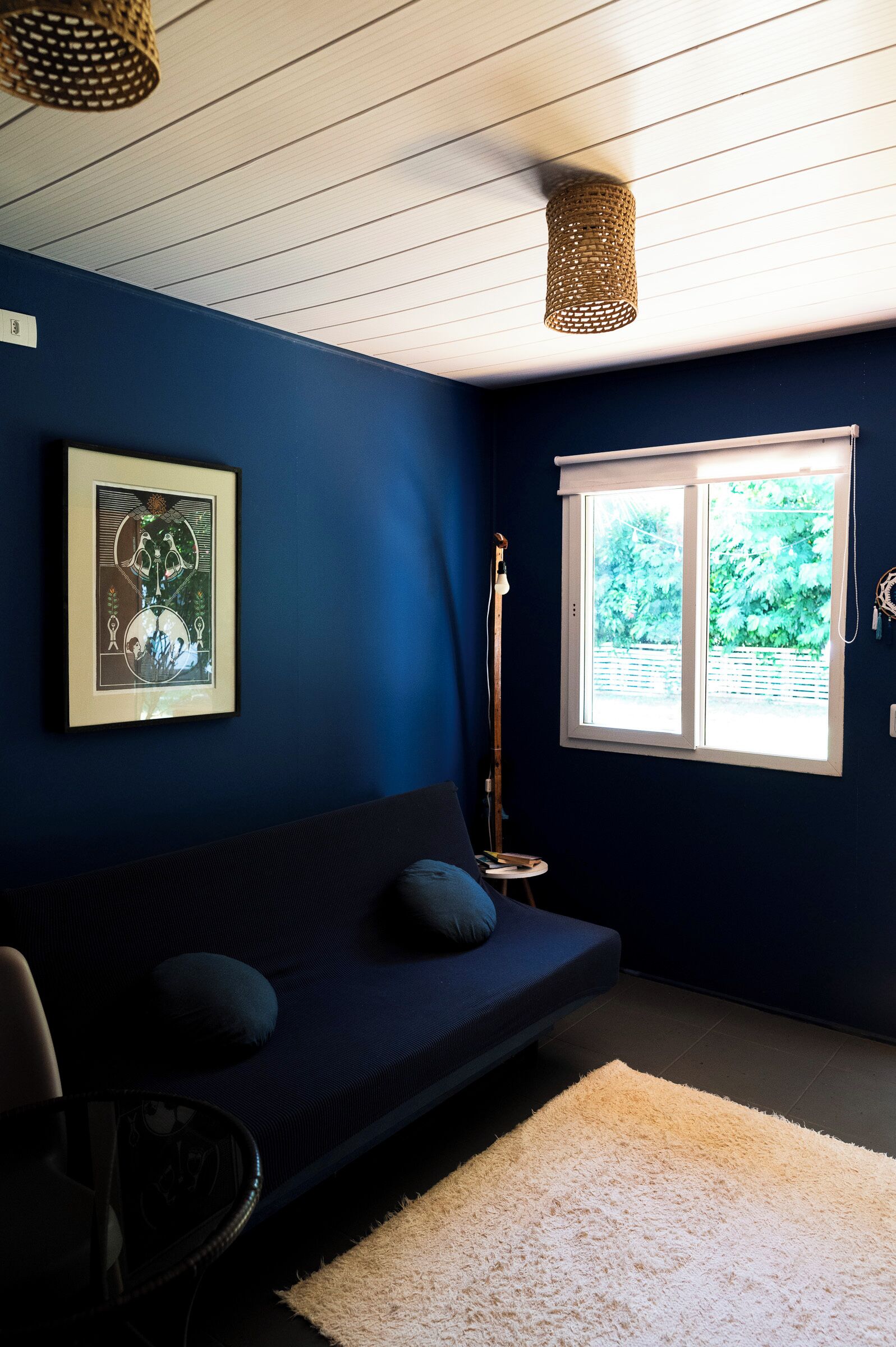 Una stanza con una parete blu scuro, un quadro incorniciato, una finestra che si affaccia sul verde e un tappeto bianco con un piccolo tavolo rotondo.