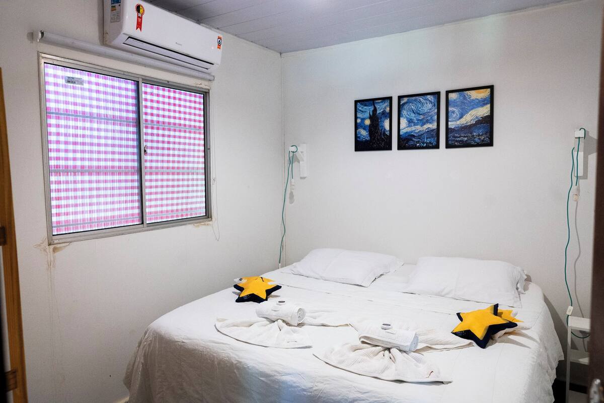 Een hotelkamer met een bed, een raam met roze geruit jaloezieën en drie schilderijen aan de muur.