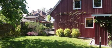A red building with a sloped roof, a small porch, and a well-maintained garden with various plants and a lawn.
