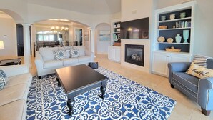 A spacious living room with a fireplace, a ceiling fan, and a dining area in the background. - Front Row Beach House 6BR/5.5 bath private pool and private beach access. (Miramar Beach)