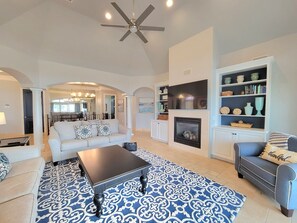 A spacious living room with a fireplace, a ceiling fan, and a dining area in the background. - Front Row Beach House 6BR/5.5 bath private pool and private beach access. (Miramar Beach)