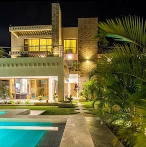 A modern two-story house with a swimming pool, stone facade, and well-lit windows.