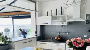 Ett modernt kök med en central köksö, vita skåp och utsikt över en vattenyta genom ett stort fönster.