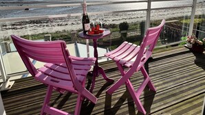 En balkong med två rosa stolar, ett litet bord med en flaska och glas, och utsikt över stranden och havet.