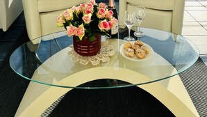 Ett rum med två vita läderfåtöljer placerade mittemot ett glasbord. På bordet står en vas med rosa blommor, en flaska champagne och en tallrik med kakor. Ett stort fönster ger utsikt över havet och en avlägsen kustlinje.