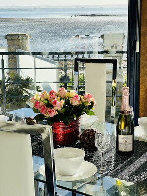 A table set with a bottle of Louis Boulot Brut, a vase of pink roses, and a glass. The table is on a balcony with a view of the sea.