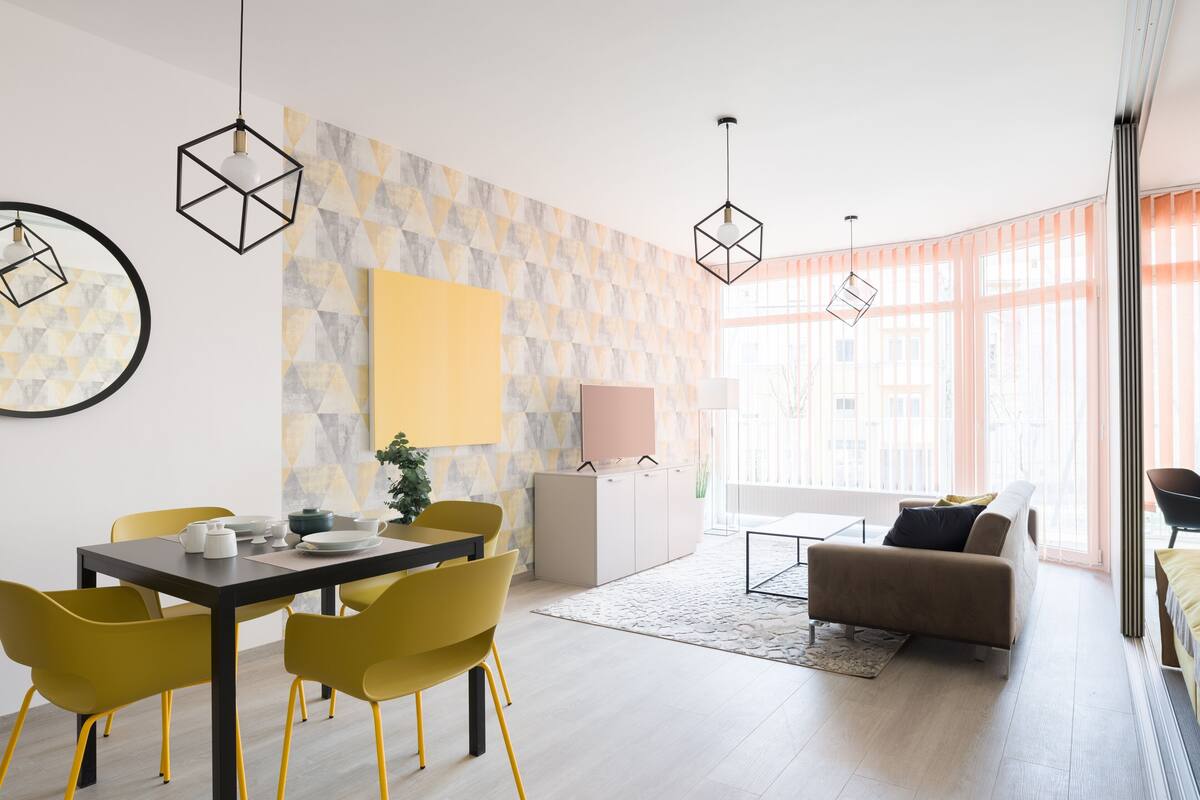 Ein modernes Wohnzimmer mit Essbereich, mit geometrischen Wandmustern, gelben Stühlen und Pendelleuchten.