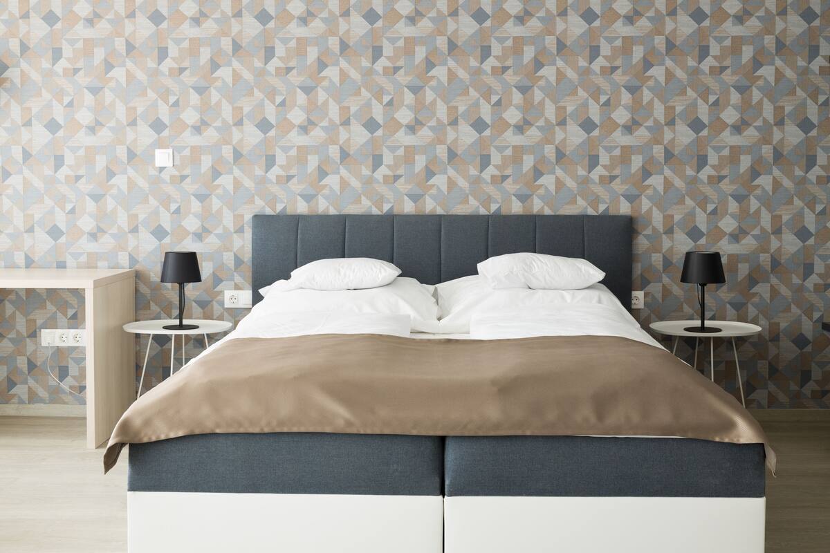 Ein modernes Schlafzimmer mit einer Wand in geometrischem Muster, einem grauen, gepolsterten Kopfteil, weißer Bettwäsche und zwei Nachttischen mit Lampen.