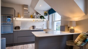 A modern kitchen with grey cabinetry, a built-in oven, a microwave, and a coffee machine. There is a window with a view of the outdoors, and a bar stool with a plaid cushion. - Howe Keld 6 Second Floor (Keswick)