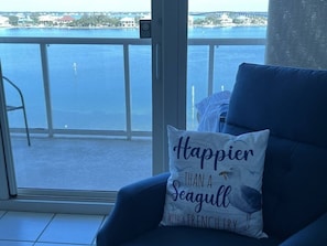 A balcony with a view of the water, a chair with a cushion, and a window with a view of the water.