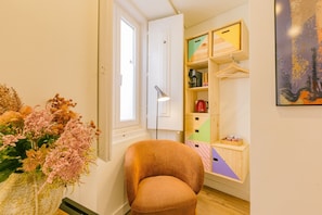 Una habitación con una silla, un florero de flores, una estantería con una tetera y un cuadro en la pared.