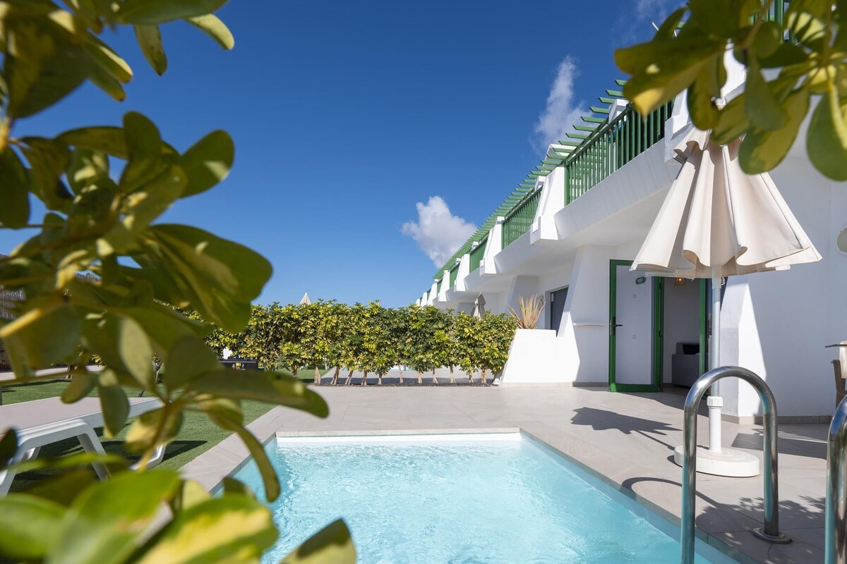 A swimming pool with a clear blue water surface, surrounded by a white building with green accents and a hedge in the background.