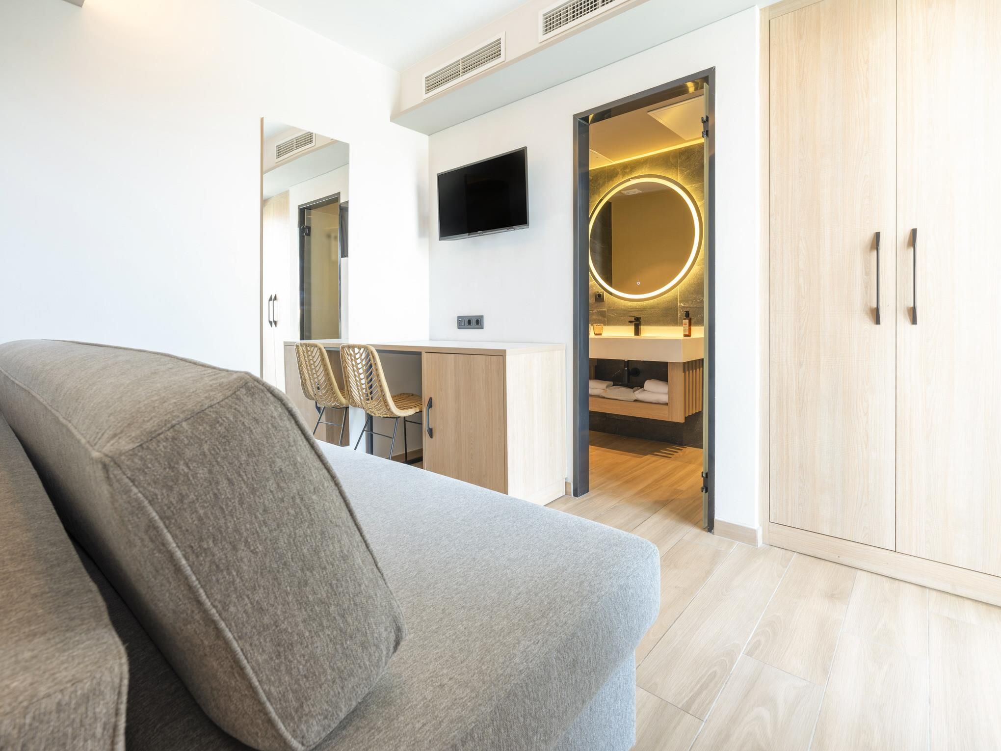 A modern hotel room with a sofa, a desk, a TV, a bathroom with a mirror, and wooden flooring.