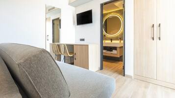 Una habitación de hotel moderna con sofá, escritorio, televisor y baño con espejo, además de suelo de madera.
