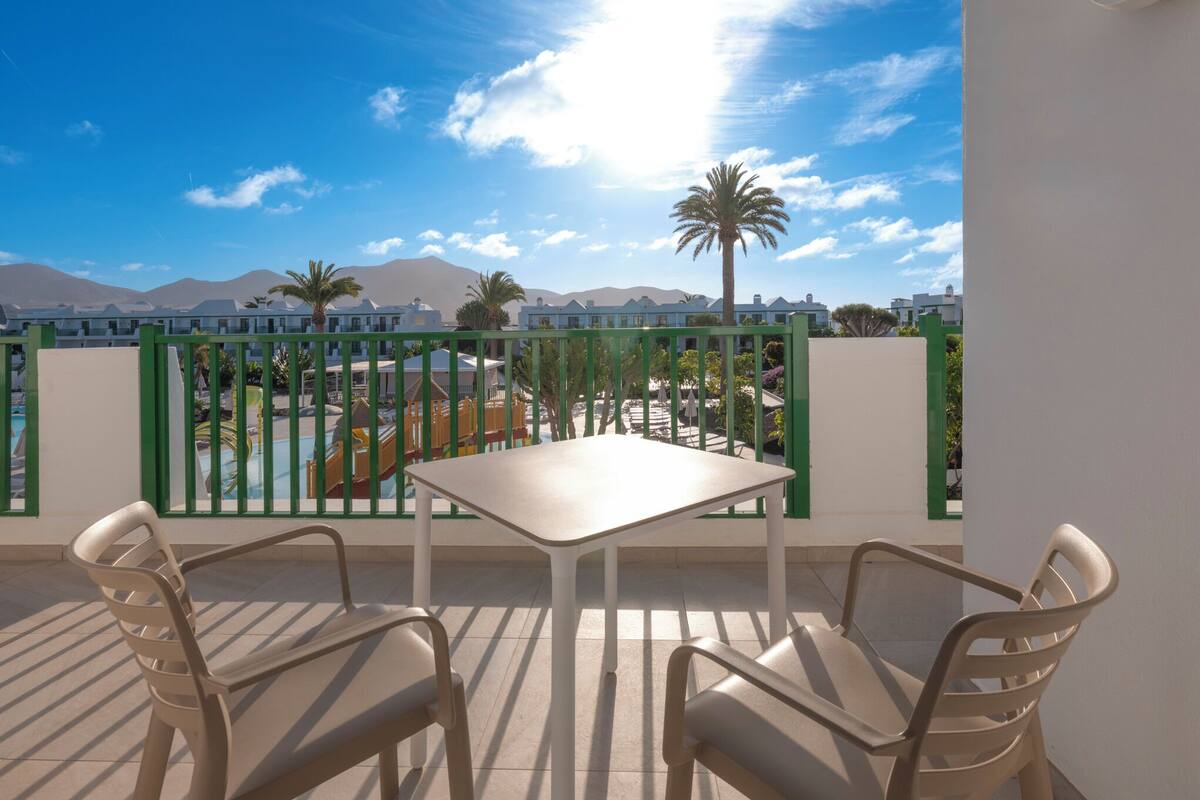 A balcony with a table and chairs, overlooking a pool area with palm trees and mountains in the background.