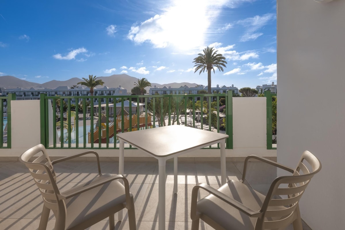 Un balcón con mesa y sillas, con vistas a una piscina y edificios, con montañas al fondo.