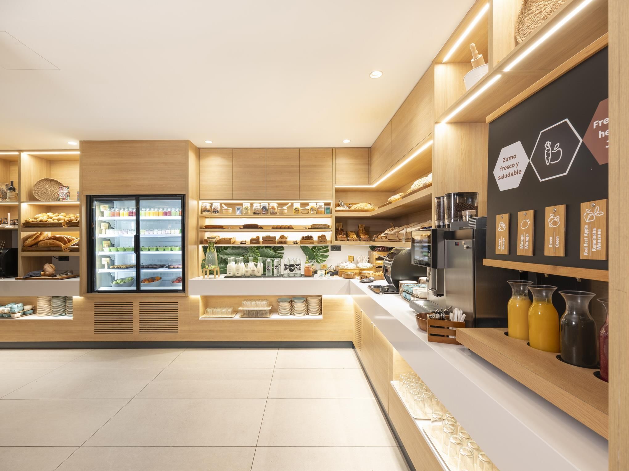 Un interior de cafetería moderna con una estación de autoservicio para bebidas, un refrigerador con productos y varios artículos alimenticios en estanterías.