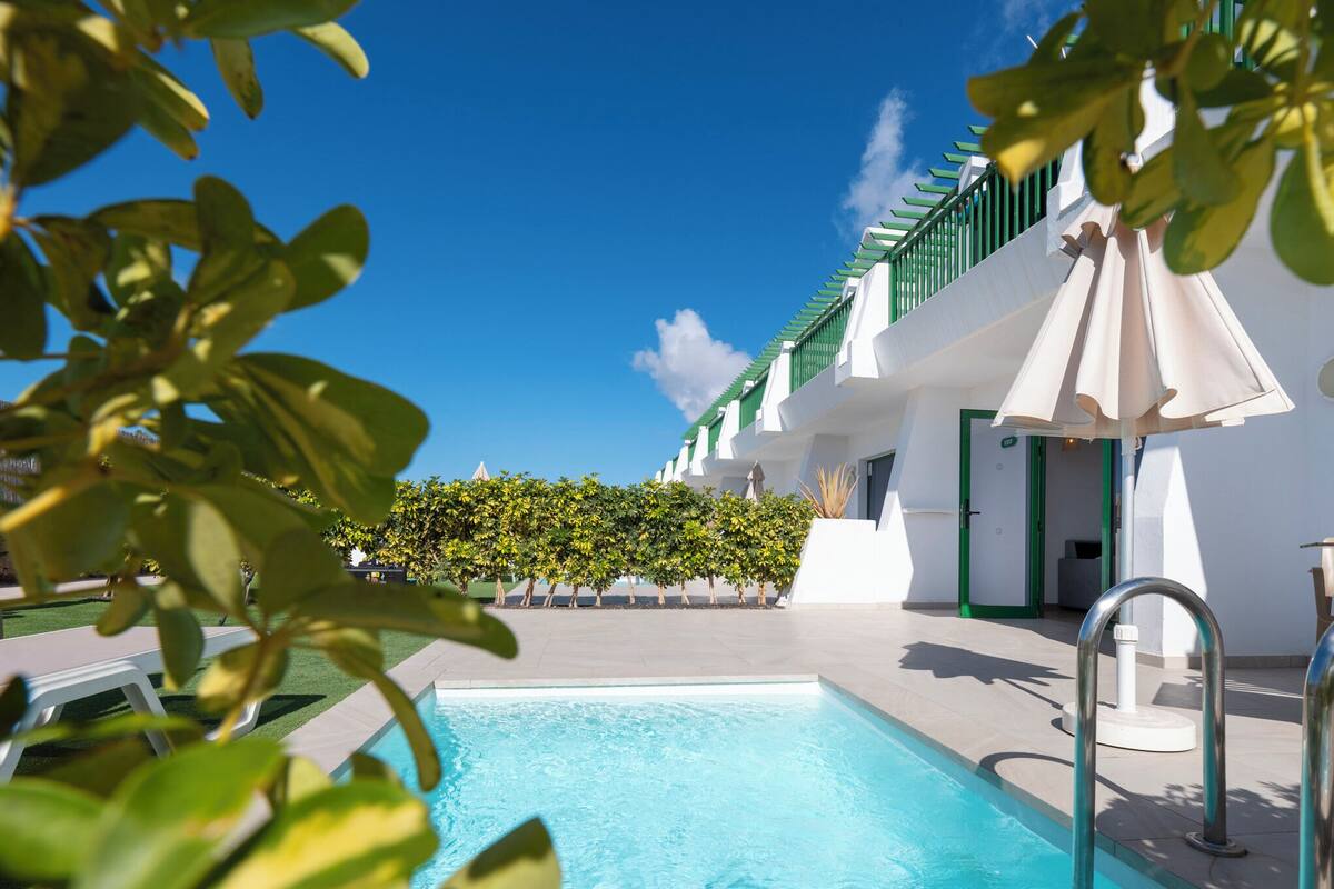 A swimming pool with a clear blue water, surrounded by a white building with green accents and a hedge.
