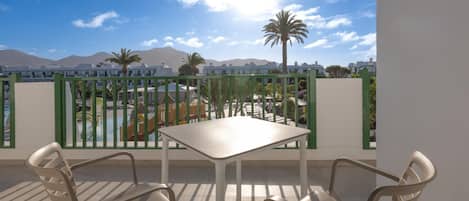 A balcony with a table and chairs, overlooking a pool and buildings with mountains in the background.