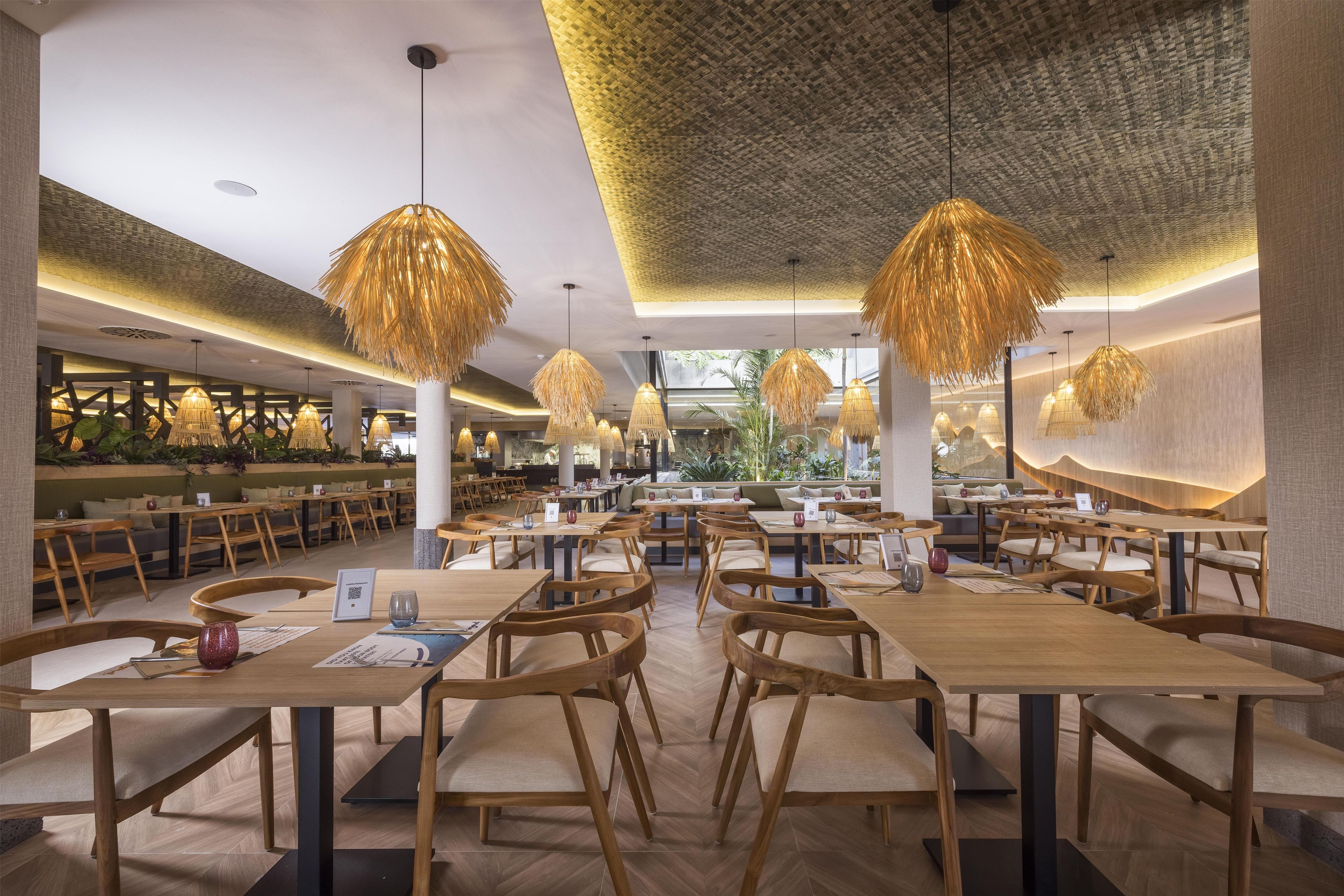 A spacious restaurant with wooden tables and chairs, hanging lamps, and a view of the outdoor area.