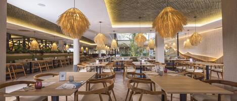 A spacious restaurant with wooden tables and chairs, hanging lamps, and a view of the outdoor area.