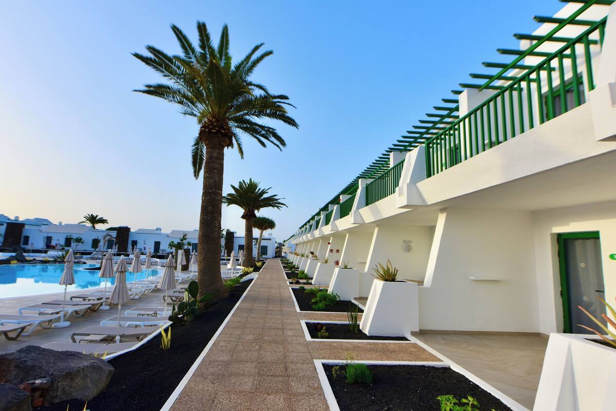 A poolside area with lounge chairs and umbrellas, a pathway lined with palm trees, and a modern building with a green railing.