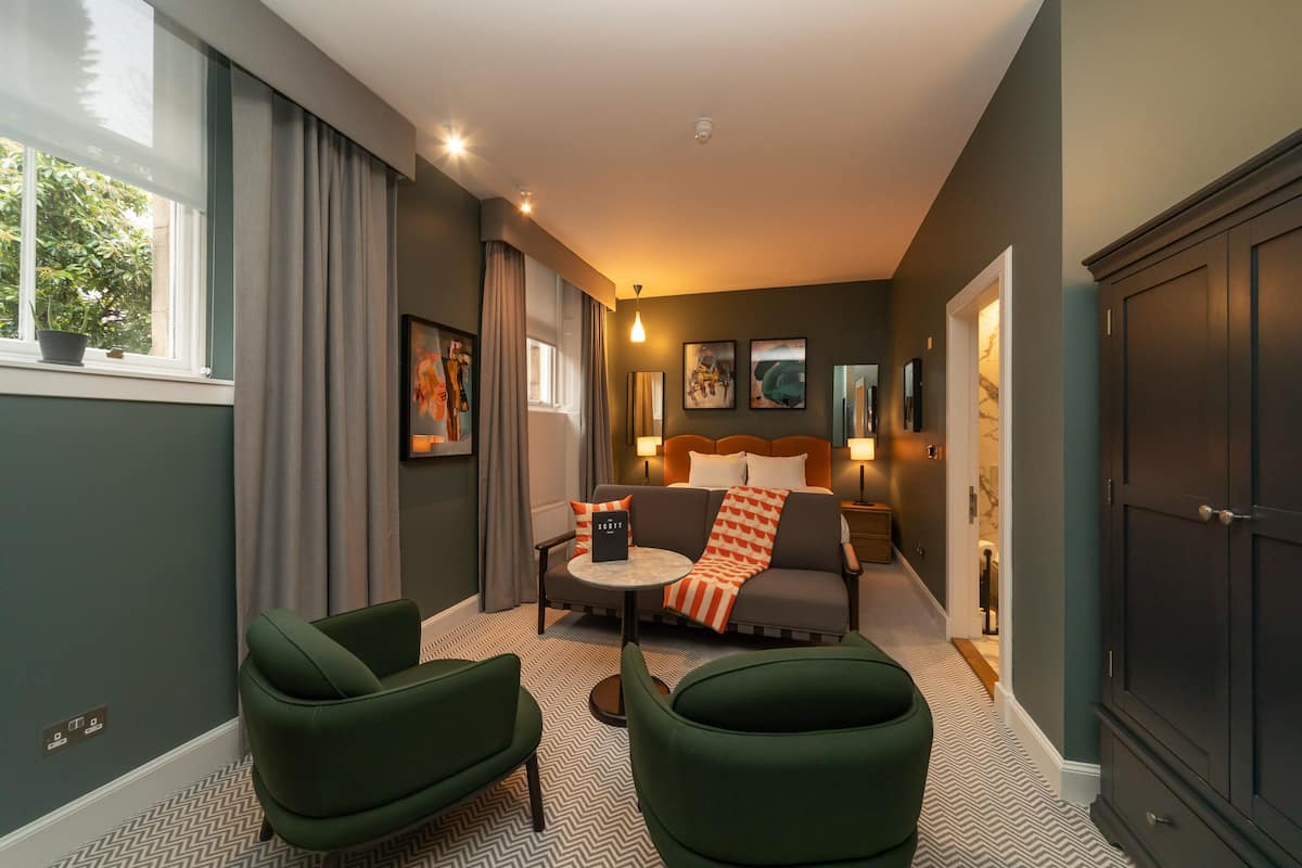 Ein modernes Hotelzimmer mit einer Couch, zwei Sesseln, einem kleinen Tisch und einem Bett mit Kopfteil. An den Wänden befinden sich Gemälde, ein Fenster mit Vorhängen und eine Tür zu einem weiteren Raum.
