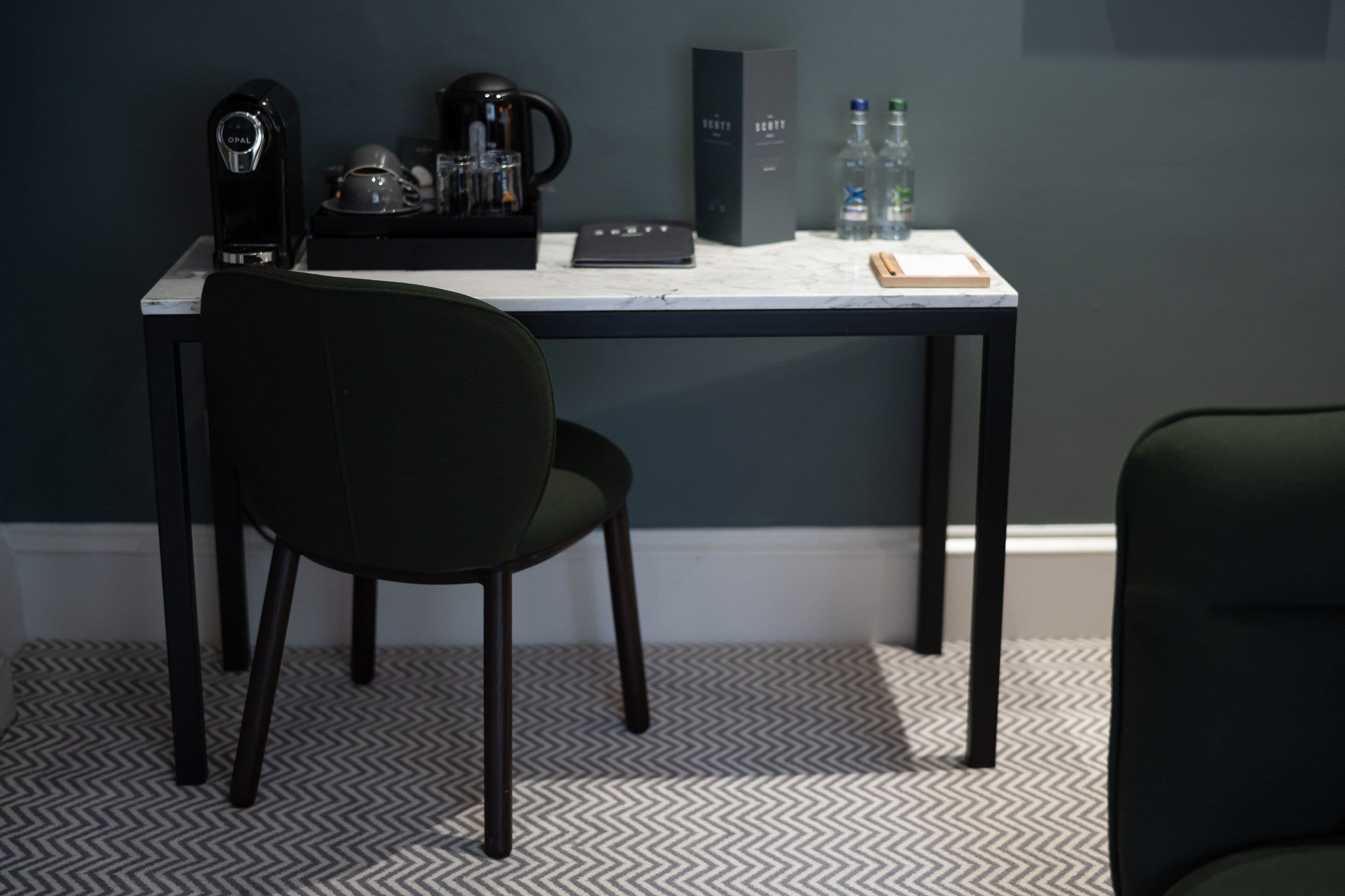 A modern hotel room with a dark green chair, a white desk with a black box, a coffee maker, and two bottles of water.