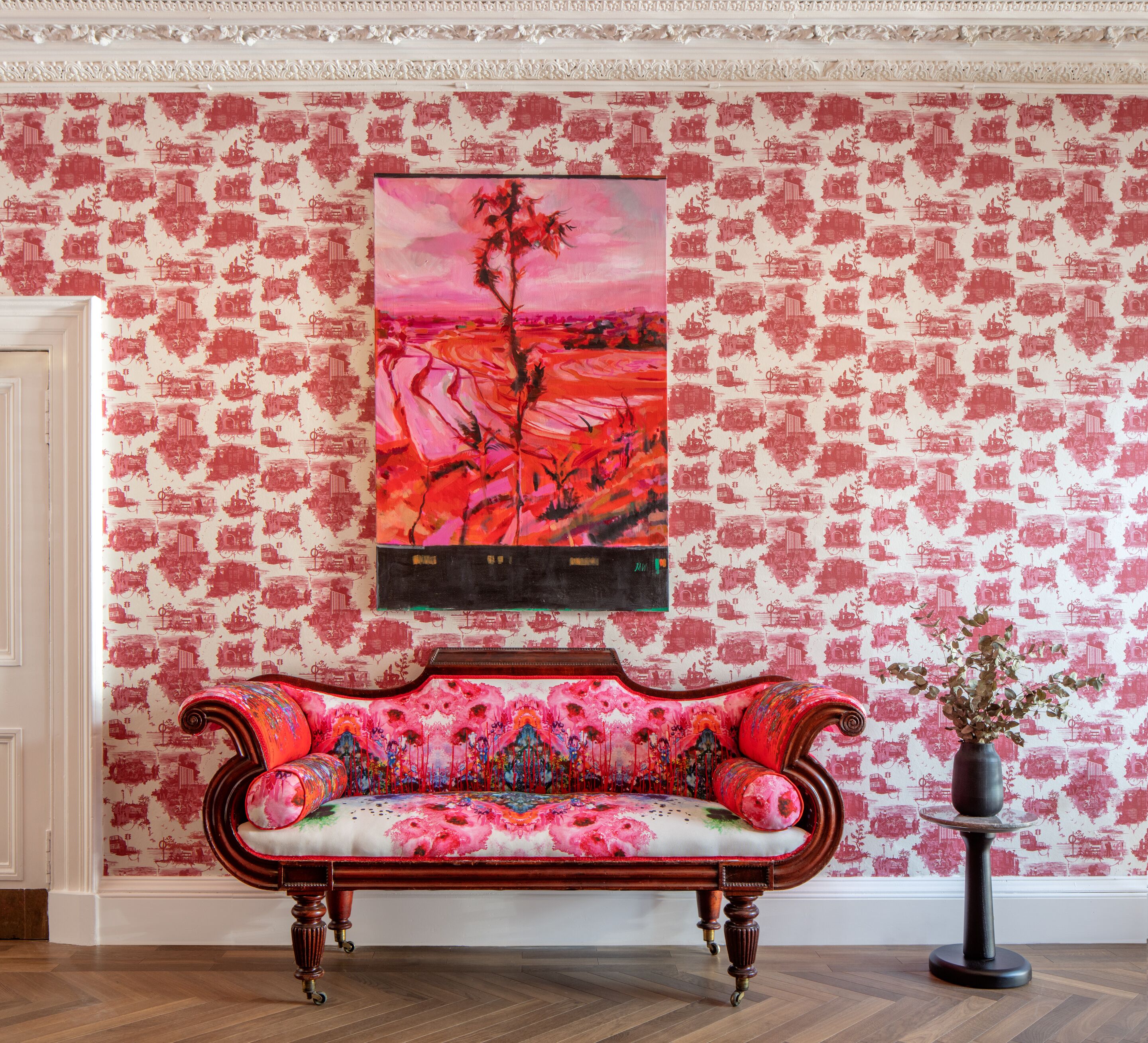 A room with a patterned wallpaper, a colorful sofa, and a painting on the wall.