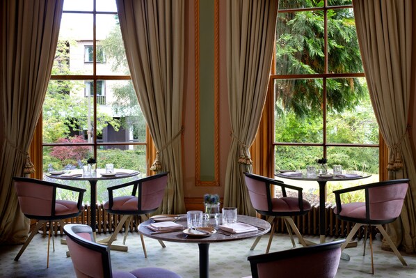 A dining area with round tables and chairs, large windows with curtains, and a view of greenery outside. - The Scott (Edinburgh)