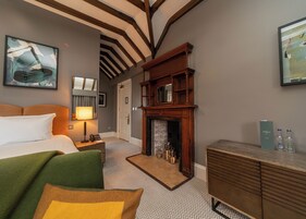 A hotel room with a bed, a green armchair, a fireplace, a nightstand with a lamp, and a framed picture on the wall.