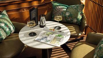 Une table ronde est dressée dans une pièce où l’on trouve des coussins à motifs et des meubles en bois. Sur la table, on aperçoit deux verres de vin, un menu et un objet décoratif.