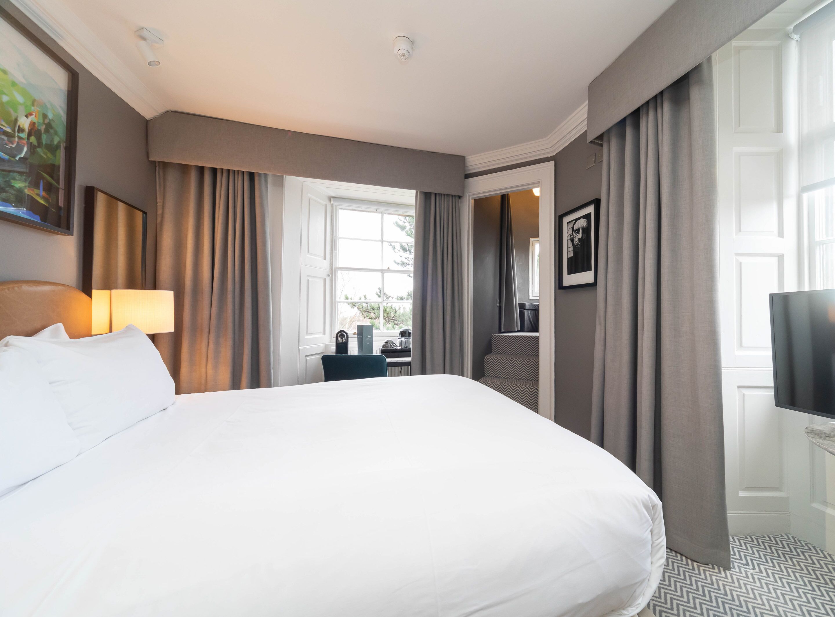 Habitación de hotel con una cama grande, un escritorio con silla, televisor y ventana con cortinas.