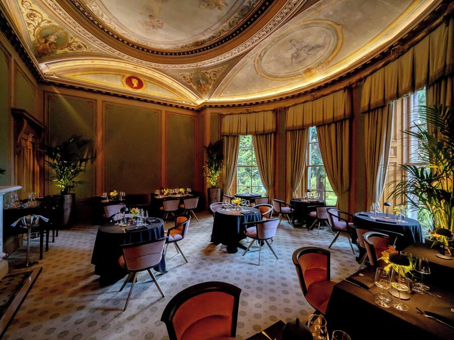 Un comedor elegante con varias mesas y sillas, una chimenea y grandes ventanales con cortinas.