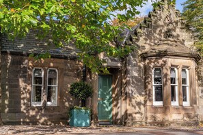A historic stone building with a green door, a window with a white frame, and a small garden area with a planter.