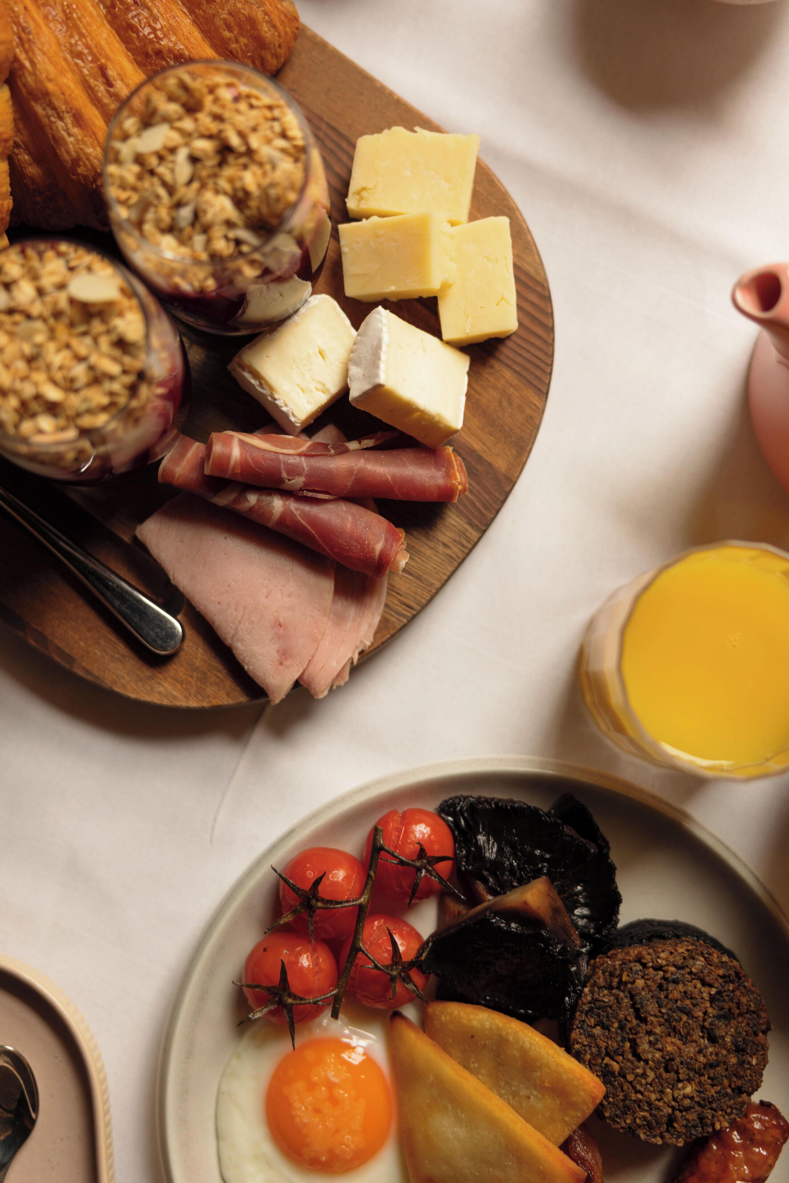 Una bandeja de desayuno con varios quesos, carnes y frutas sobre una tabla de madera.