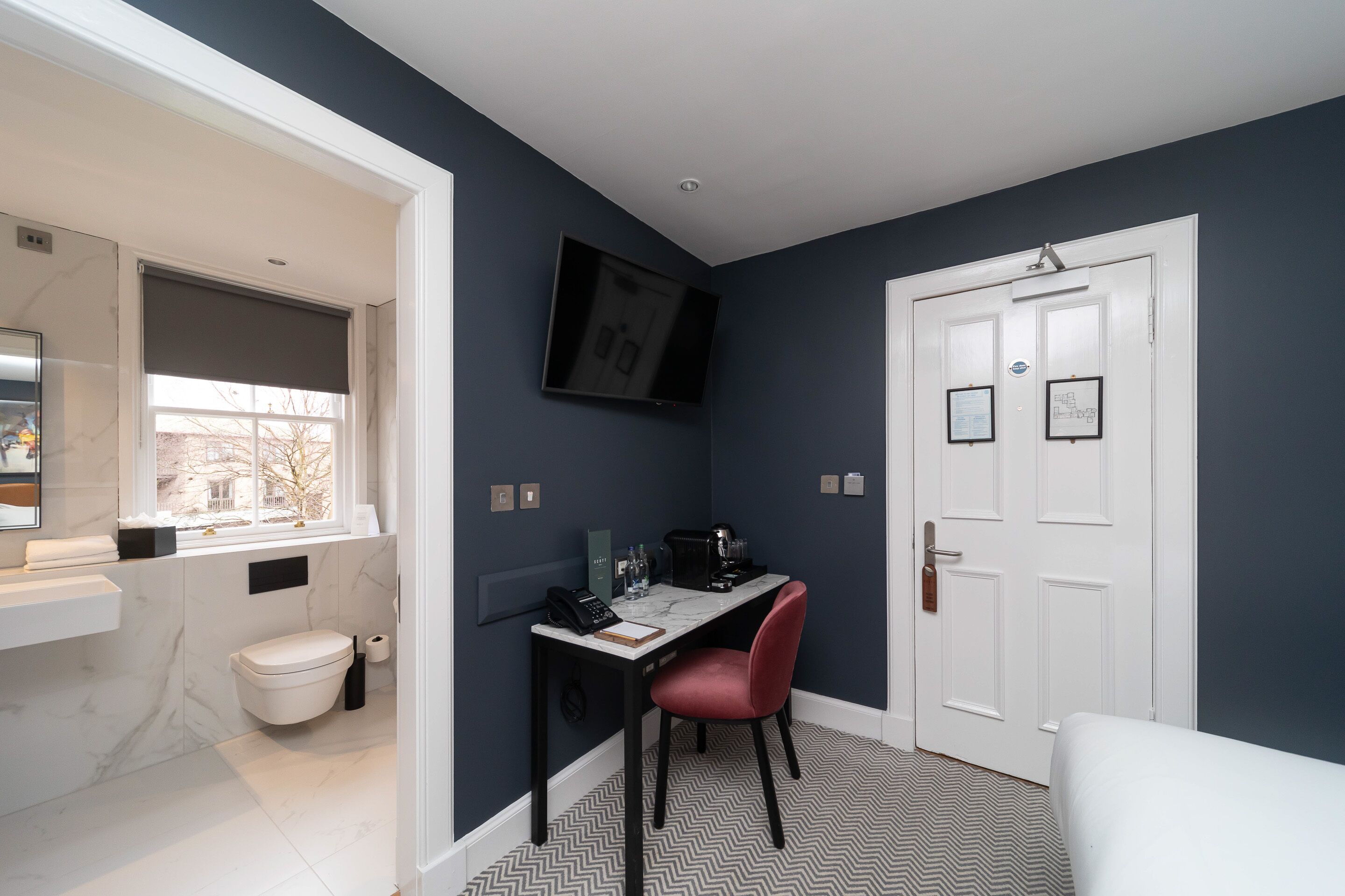 A modern bathroom with a white toilet, a sink, and a window. There is a desk with a chair and a flat-screen TV mounted on the wall.