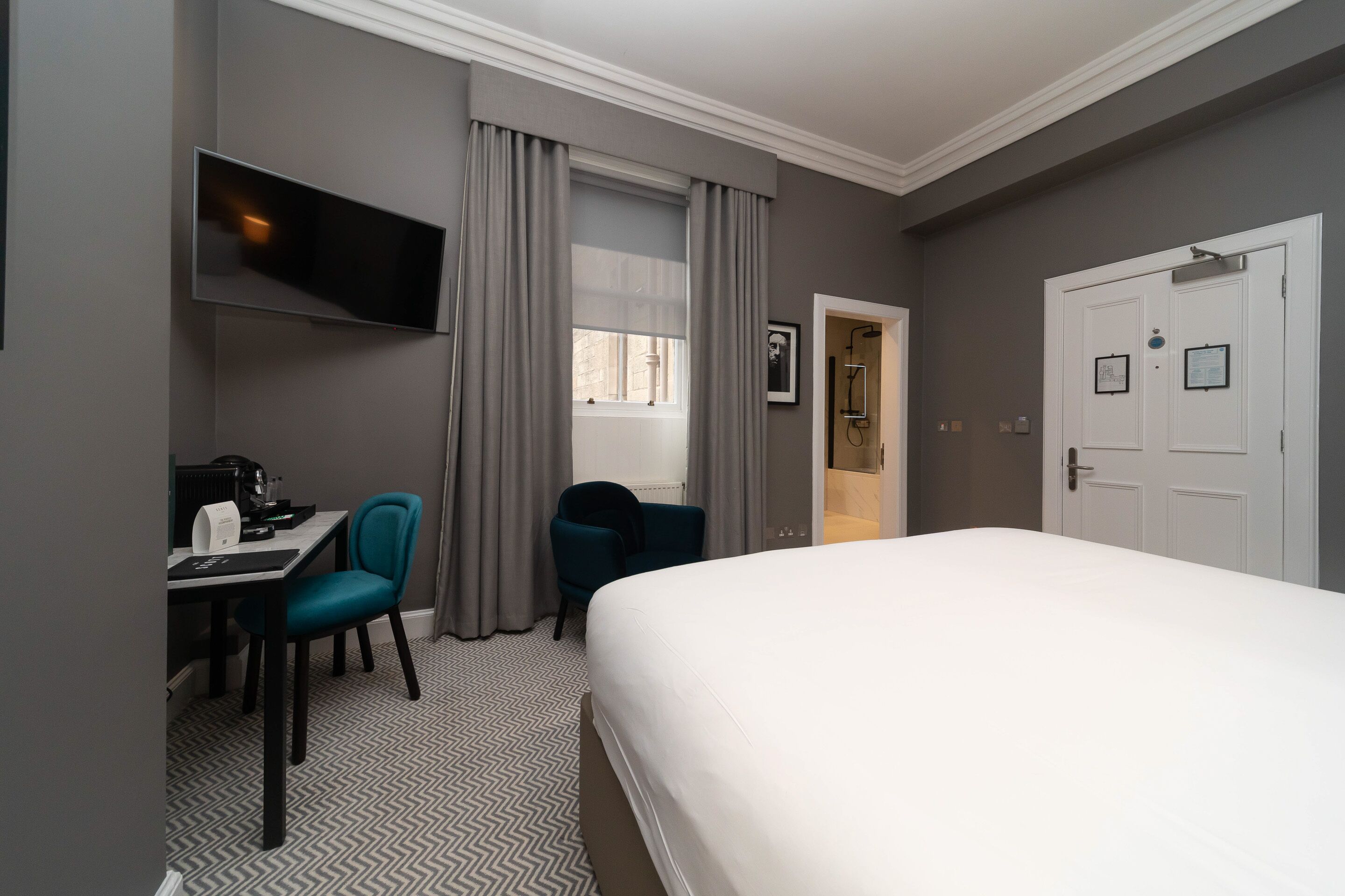 Una habitación de hotel con una cama, un escritorio con una silla, un televisor montado en la pared y una puerta con un letrero.
