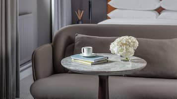 Habitación de hotel con sofá, una mesita redonda con un libro y una taza, cama con ropa blanca y dos cuadros en la pared.