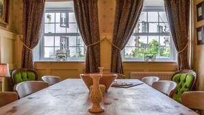 Ein Essbereich mit Holztisch, grünen Polsterstühlen und Blick auf Gebäude durch große Fenster mit gemusterten Vorhängen.