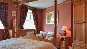 Ein Schlafzimmer mit einem Himmelbett, einem Nachttisch mit Lampe, einem Schminktisch und einem Fenster mit Vorhängen.