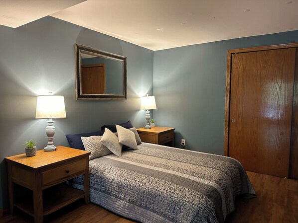 Een slaapkamer met een bed, twee nachtkastjes, een spiegel, een houten deur en een bijzettafeltje met een plant.