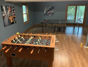 Een kamer met een tafelvoetbaltafel, een pingpongtafel en twee ramen met uitzicht op groen.