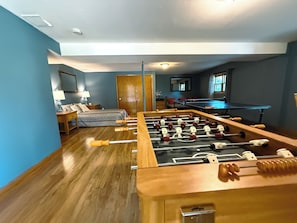 A room with a foosball table, a bed, a desk with a lamp, and a window.