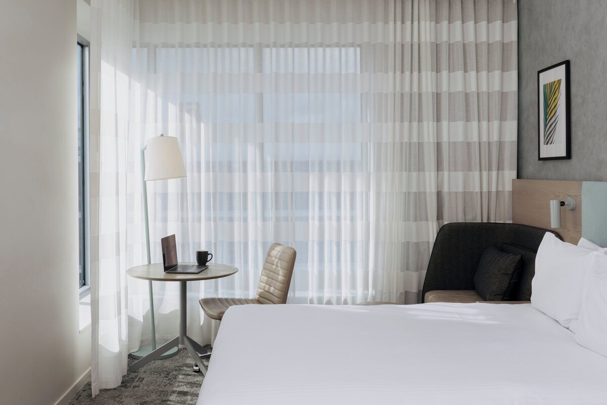 Habitación de hotel con una cama, dos sillas, una mesita con una computadora portátil y una taza, y una ventana con cortinas translúcidas.